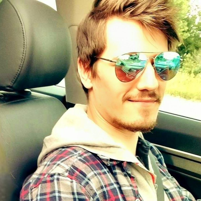 A person wearing reflective sunglasses, a flannel shirt, and a hoodie sits in a car, smiling at the camera.