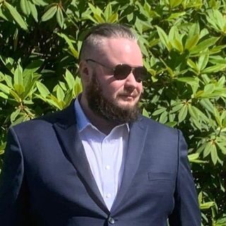 Man with a beard and sunglasses in a blue suit stands outdoors in front of green foliage.